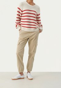Osoba ubrana w sweter w kremowo-czerwone paski, beżowe spodnie cargo z elastycznymi mankietami oraz białe sneakersy na gumowej podeszwie stoi.