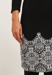 Main reposant à côté d'une robe noire avec un motif floral blanc et ornemental complexe près de l'ourlet, sur un fond neutre.