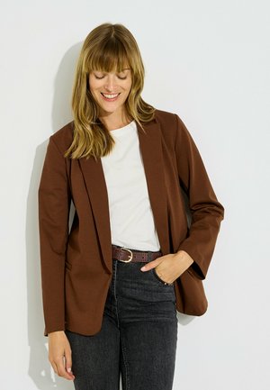 Mujer sonriendo con los ojos cerrados, vistiendo una blazer marrón, camisa blanca y vaqueros negros con la mano en el bolsillo, sobre un fondo blanco.