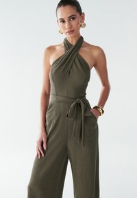 Combinaison verte olive en tissu texturé, décolleté halter, détail de taille à nouer et coupe ample. Boucles d'oreilles et bracelet en or accentuent le look.
