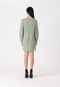Robe en tricot vert avec une coupe décontractée, manches longues et encolure large. Tissu texturé, se terminant au-dessus du genou, associée à des chaussures noires.