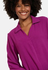Chemise longue à manches violettes avec col, présentant un tissu texturé et un design en col en V. Le mannequin sourit, mettant en valeur le haut.