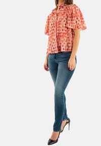 Donna che indossa una blusa corta a maniche corte rossa con un motivo, jeans aderenti blu e scarpe con tacco alte nere, in piedi davanti a uno sfondo bianco.