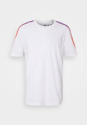 Homme portant une chemise blanche à manches courtes avec des rayures orange, un jean bleu clair et des baskets montantes vert foncé, debout devant un fond blanc.