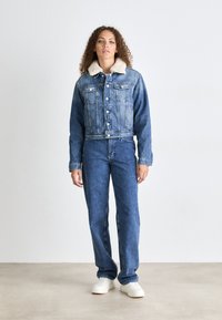 Chaqueta de denim azul claro con un cuello de borrego crema, bolsillos frontales y cierre de botones, combinada con unos jeans rectos a juego y zapatillas blancas.