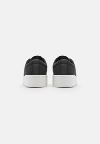 Zapatillas de lona negras con suela blanca plana, que cuentan con un cuello acolchado, refuerzo en la parte trasera y un diseño minimalista.