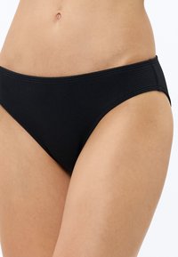 Zbliżenie osoby noszącej czarne, teksturowane majtki od bikini, skupiające się na talii, biodrach i górnej części ud na jednolitym tle.