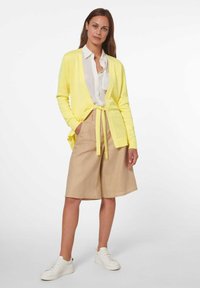 Geel gebreide cardigan met open voorkant en strik taille, gecombineerd met beige shorts en witte sneakers. Gladde textuur, casual ontwerp.