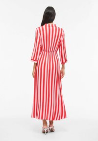 Femme de dos portant une longue robe à rayures verticales rouges et roses et des sandales blanches à talons sur un fond blanc.