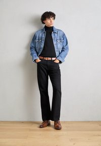 Veste en denim bleu clair, col roulé noir, pantalon noir, chaussures en cuir marron avec lacets, ceinture contrastante et accents texturés.