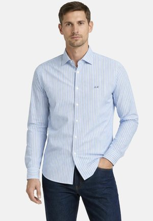 Hombre vestido con camisa de botones de rayas verticales blancas y azul claro, con vaqueros oscuros, de pie con una mano en el bolsillo.