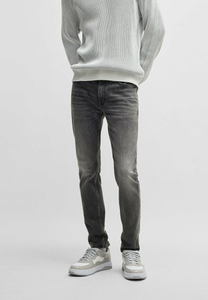 Person iført falmede sorte slim-fit jeans, en hvid ribbet sweater og hvide og grå sneakers, der står mod en ensfarvet baggrund.