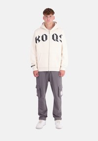 Cream zip-up hoodie with black print "KO QS" in front, paired with gray cargo pants featuring pockets and a white label. White sneakers worn.