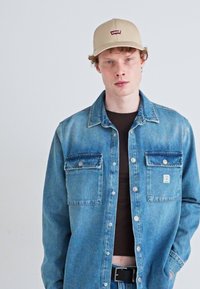 Lichtblauwe denimjas met twee voorzakken, zilveren drukknoopjes en een klein logopatch. Gecombineerd met een donkerbruine shirt en een beige pet.