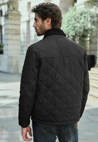 Black quilted jacket with a collar, featuring a diamond pattern, and side pockets. Worn with dark jeans, viewed from the back.