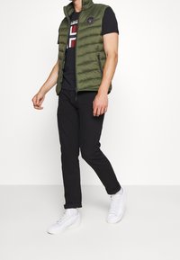 Groene gewatteerde vest met opstaande kraag, ritssluiting en logopatch, gecombineerd met een zwart graphic t-shirt en zwarte broek, afgewerkt met witte sneakers.