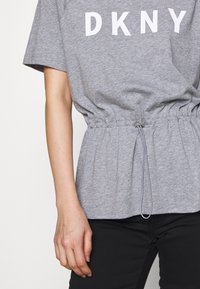Camiseta gris con gran texto blanco "DKNY", con cintura ceñida y cordón ajustable. Tejido suave, corte relajado, mangas cortas.