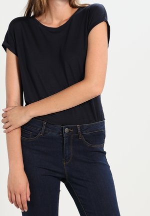 Woman wearing a plain black short-sleeve shirt and dark blue jeans with one arm crossed over her waist.