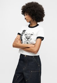 Γυναίκα με σγουρά μαλλιά που φοράει λευκό t-shirt με μαύρη περιμετρική ταινία και σχέδιο από καρτούν λαγουδάκι, συνδυασμένο με σκουρόχρωμο τζιν παντελόνι με εμπρόσθιες τσέπες.