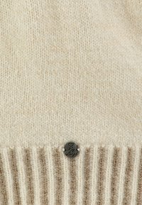 Tissu tricoté couleur crème avec bord inférieur côtelé et petite plaque métallique ronde gravée d'un cœur et d'un texte en écriture cursive.
