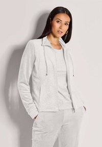 Vrouw met lang, donker haar in een witte, gemusterde ritsjack en bijpassende broeken, staat met haar handen in de zakken tegen een beige muur.