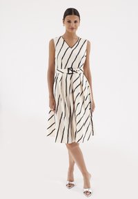 Sukienka bez rękawów w kolorze kremowym z czarnymi diagonalnymi paskami, dekolt w kształcie litery V, dopasowana talia z paskiem i rozkloszowanym dołem. Gładka struktura materiału, luźny krój.