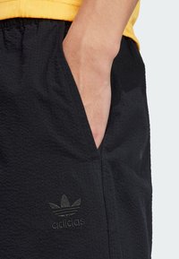 Black athletic shorts in a textured fabric, featuring a side pocket and embossed Adidas logo. Elastic waistband.