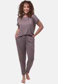 Un elegante jumpsuit marrone chiaro realizzato in un tessuto morbido, con maniche corte, vestibilità rilassata, tasche laterali e gambe affusolate. Il modello si trova in piedi con le mani in tasca.