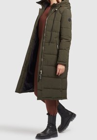 Lang, olivenfarvet pufferjakke med lynlås og trykknaplukning, samlet hætte, med quiltet tekstur og sidelommer. Sorte støvler synlige.