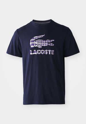 Camiseta de manga corta azul marino con un logo de cocodrilo en cuadros morados y blancos y el texto "LACOSTE" en la parte frontal.