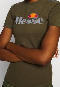 Camiseta de algodón verde oliva con mangas cortas, que presenta un gran logo "Alesse" en gris y un gráfico colorido de sol arriba.