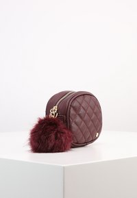 Round burgundy faux leather bag with diamond quilt pattern, gold zipper, and a burgundy fur pom-pom attached. Smooth texture, compact design.