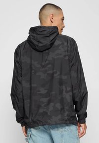 Zwarte camouflagejas met capuchon, lichtgewicht materiaal, elastische manchetten en een losse pasvorm, weergegeven van achteren tegen een effen achtergrond.