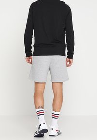 Schwarzes Langarmshirt kombiniert mit grauen Shorts. Weiße Socken mit roten und blauen Streifen. Sportschuhe mit schwarz-weißen Akzenten.