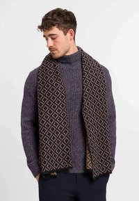 Uomo che indossa un maglione a collo alto bordato viola e una sciarpa nera con un motivo geometrico a rombi marroni, mani in tasca, che guarda in basso.