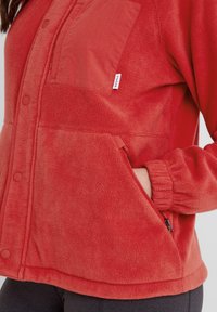Red fleece jacket with a smooth fabric pocket, snap buttons, elastic cuffs, and a side zip pocket. The texture appears soft and warm.
