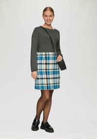Grijze top met lange mouwen gecombineerd met een blauw, zwart en wit geruit rokje. Het model houdt een kleine zwarte tas vast en draagt zwarte instap schoenen.