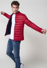 Rød polstret jakke med høj krave, frontlynlås og sidelommer, båret over en hvid t-shirt og parret med blå jeans og hvide sko.