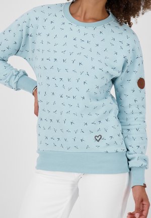 Femme portant un sweat-shirt bleu clair avec de petits motifs d’oiseaux, un emblème en forme de cœur près de l’ourlet, des manches retroussées et un pantalon blanc.