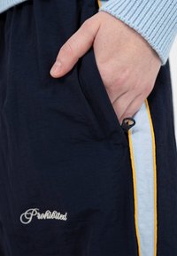 Ranka kišeniuose tamsiai mėlynų kelnių su šoniniu užtrauktuku ir geltonai-mėlyna juostele, dėvima kartu su šviesiai mėlynu, raukšlėtu rankogaliu.