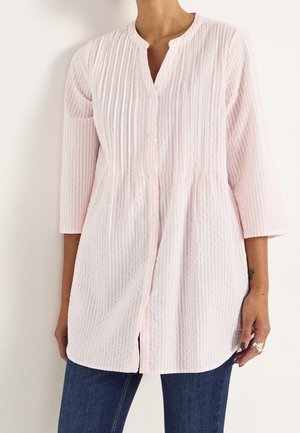 Persona vistiendo una túnica de botones de rayas verticales en rosa claro con mangas 3/4 y jeans azul oscuro, de pie contra un fondo blanco.