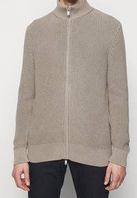 Beige stickad ziptröja med hög krage, strukturerad väv och ribbade muddar och fåll. Har en silverfärgad dragkedja och en avslappnad passform.