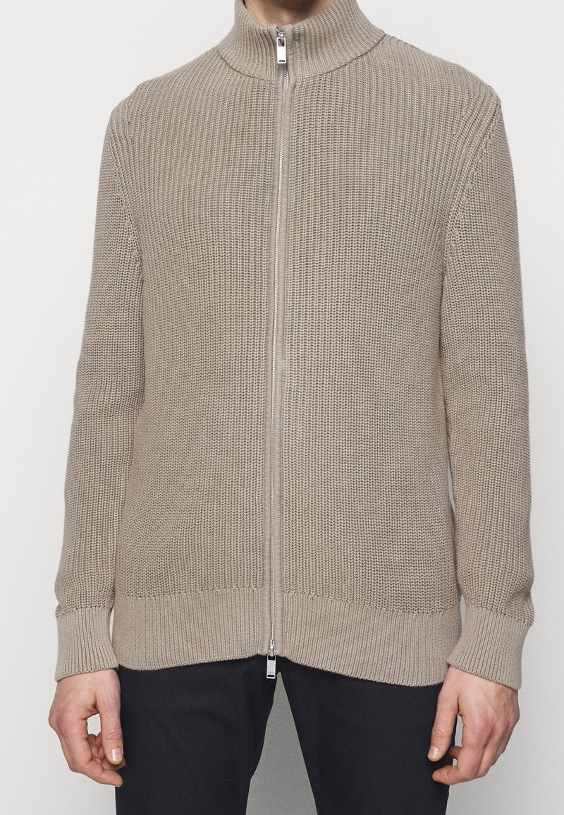 Beige stickad ziptröja med hög krage, strukturerad väv och ribbade muddar och fåll. Har en silverfärgad dragkedja och en avslappnad passform.