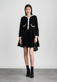 Junge Frau in einer schwarz-weißen, geknöpften Jacke, mit einem schwarzen ausgestellten Rock, schwarzen Socken, schwarzen Schuhen und einer kleinen schwarzen Handtasche.