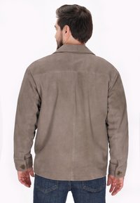 Jachetă din suede maro cu guler de cămașă, mâneci lungi și manșete cu nasturi. Are o textură netedă și un design relaxat.