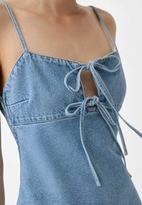 Svetle modré denimové šaty s tenkými ramínky, se středovým zavazováním, malým kapsičkovým detailem a mírně A-liniovým střihem.