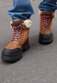 Bottines en daim et cuir marron avec semelles en caoutchouc noir, lacets à motifs rouges et noirs, et bordure en fourrure blanche autour du col.