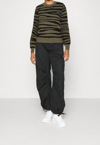 Olivgrüner Pullover mit schwarzen Zebra-Streifen, kombiniert mit lässig sitzenden schwarzen Cargo-Hosen und weißen Sneakers. Weicher Stoff und lockere Passform.