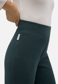Leggings de talle alto en color azul verdoso, con tejido texturizado y una etiqueta de marca en la cinturilla. Su diseño ajustado se adapta al contorno del cuerpo.