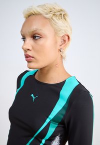 Svart tröja med långa ärmar som har tealfärgade accentremsor och en Puma-logotyp. Slät textur med en figurnära design och en hjärt-tatuering på nacken.